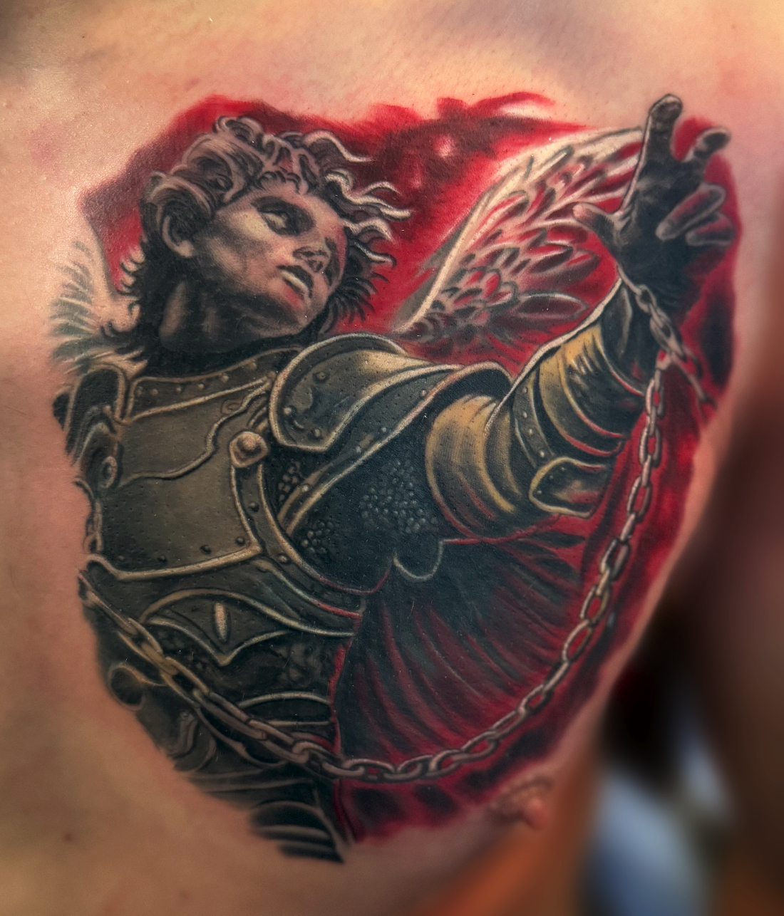 Colour realism chest tattoo of an armoured angel in chains against a red background