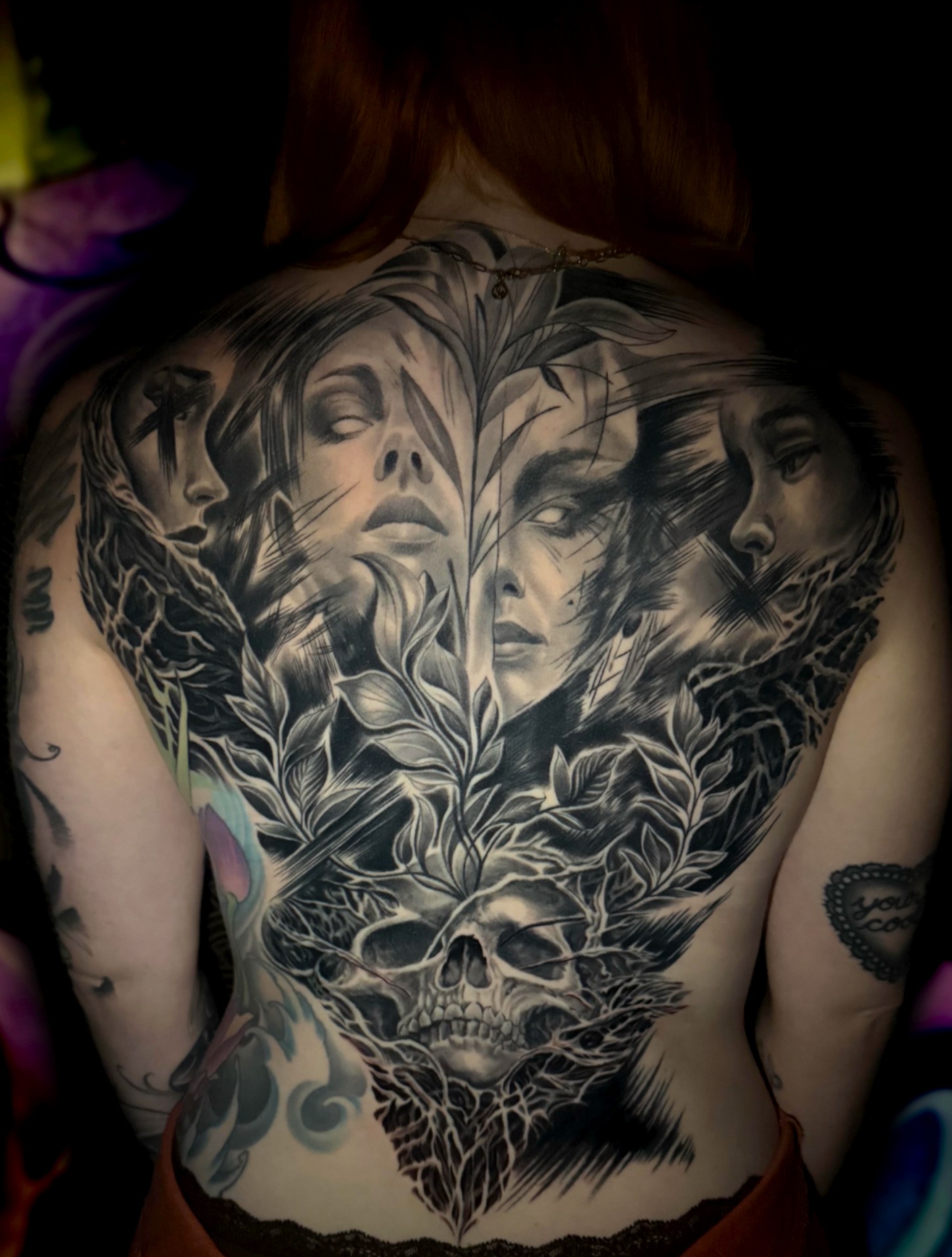 Black and grey full back piece of four women's faces among botanical leaves above a skull and roots