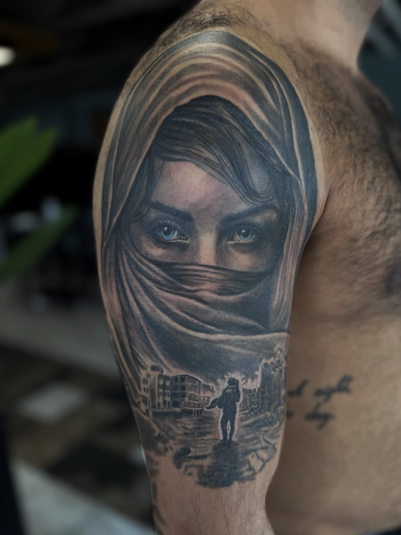 Black and grey realism shoulder tattoo of a veiled woman with striking blue eyes above a ruined cityscape